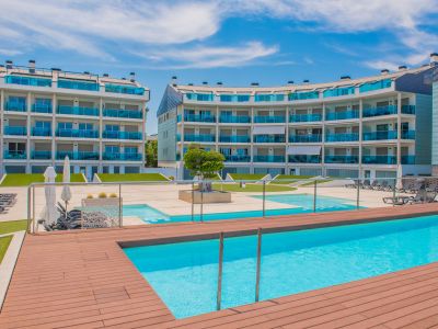 La zona de piscina comunitaria presenta dos piscinas de agua azul cristalina, rodeadas de tarima de madera, tumbonas y césped artificial, frente a edificios de apartamentos modernos.