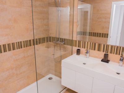Moderno cuarto de baño con ducha a ras de suelo y mampara de cristal. Destaca el lavabo doble y los azulejos beige con franja decorativa de mosaico.