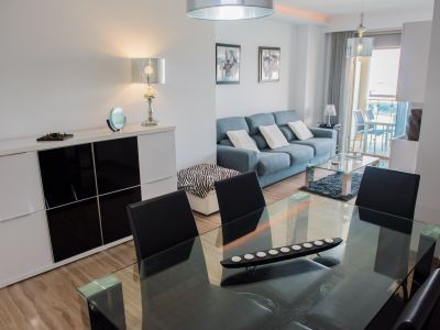 Salón-comedor de concepto abierto con mesa de cristal, sillas negras y sofá azul. El suelo de baldosas y un aparador blanco moderno complementan el espacio, con acceso a la terraza.