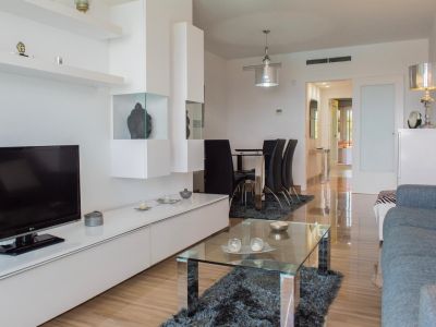 Salón-comedor moderno con mueble modular blanco, mesa de centro de cristal sobre alfombra oscura y conjunto de comedor con sillas negras. Suelos pulidos y aire acondicionado integrado.