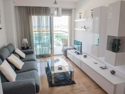 Salón moderno con sofá seccional gris y mesa de centro de cristal. Puertas correderas de vidrio dan a una terraza con vistas a la piscina y al mar. Suelos de mármol pulido.