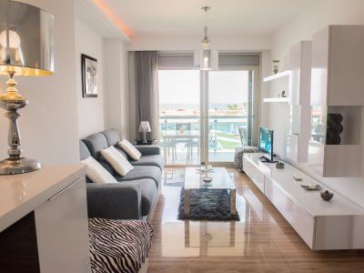Salón moderno con suelos de mármol pulido, acceso a terraza con vistas a la piscina comunitaria y al mar. Destacan sofá gris y muebles blancos contemporáneos.