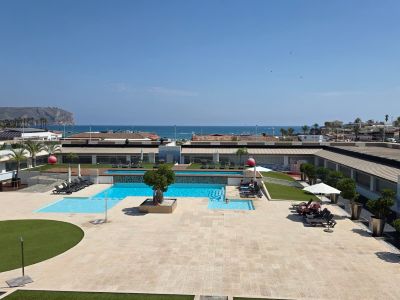 Vista aérea de la amplia zona de piscinas comunitarias con terrazas pavimentadas, césped artificial y tumbonas, en primera línea de mar con acantilado al fondo.