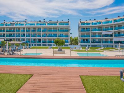 Vista de la piscina comunitaria con tarima de madera y césped artificial, flanqueada por modernos edificios residenciales con balcones de cristal azul.