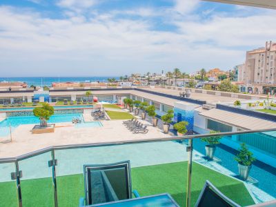 Vista elevada desde la terraza con barandilla de cristal, mostrando la piscina comunitaria, zonas de césped artificial y el mar Mediterráneo a lo lejos.