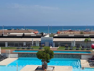 Vista panorámica de las piscinas multinivel del complejo, con tumbonas, césped artificial y vistas directas al mar Mediterráneo y la costa lejana.