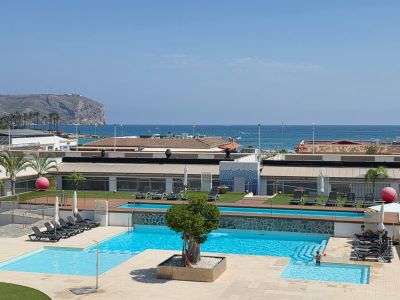 Vista panorámica del área de piscinas de varios niveles con un árbol central, rodeada de tumbonas y césped artificial, con el mar Mediterráneo y acantilados lejanos de fondo.