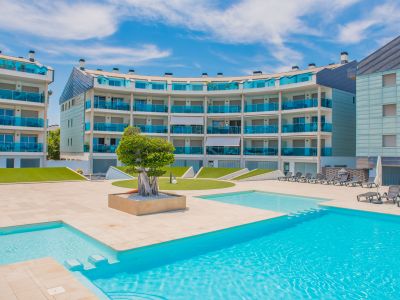Zona de piscina comunitaria con pavimento claro, tumbonas y edificios residenciales modernos con balcones curvos de cristal azul.