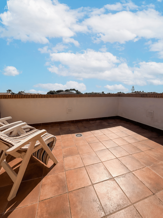 Amplia terraza exterior con suelo de baldosas de terracota, un murete blanco con remate de ladrillo y dos sillas plegables de madera, bajo un cielo azul con nubes. Amplia terraza exterior con suelo de baldosas de terracota, un murete blanco con remate de ladrillo y dos sillas plegables de madera, bajo un cielo azul con nubes.