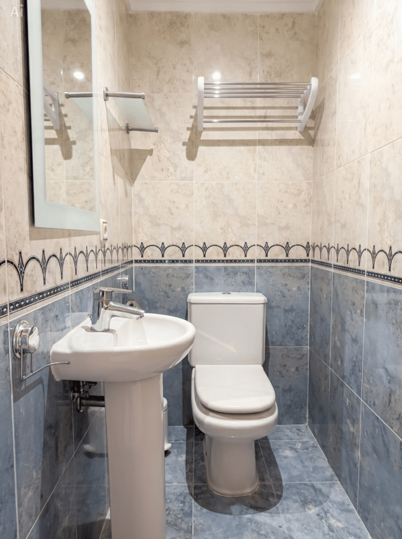 Aseo de cortesía compacto con lavabo de pedestal, inodoro y paredes alicatadas en dos tonos de azul y beige con cenefa decorativa. Incluye espejo y toalleros. Aseo de cortesía compacto con lavabo de pedestal, inodoro y paredes alicatadas en dos tonos de azul y beige con cenefa decorativa. Incluye espejo y toalleros.