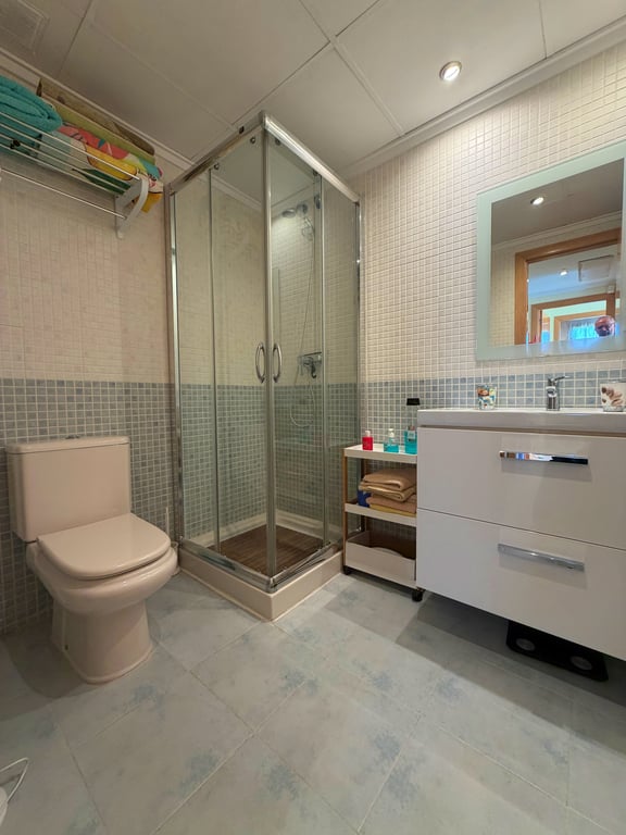 Baño funcional con ducha de cristal, inodoro en tono beige y un mueble de lavabo blanco con espejo iluminado. Las paredes combinan mosaicos claros y azules, complementados por el suelo de baldosas a juego. Baño funcional con ducha de cristal, inodoro en tono beige y un mueble de lavabo blanco con espejo iluminado. Las paredes combinan mosaicos claros y azules, complementados por el suelo de baldosas a juego.