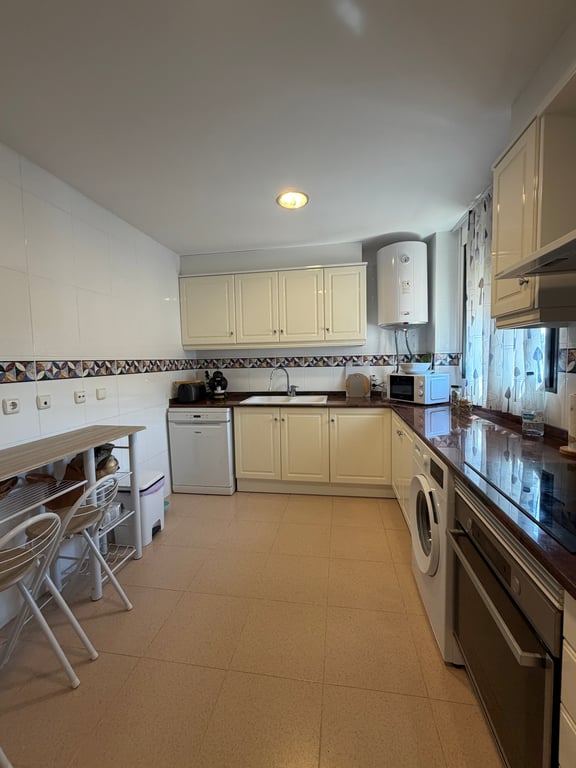Cocina cerrada con armarios color crema, encimeras oscuras y electrodomésticos integrados. Incluye una barra de desayuno de madera y suelo de baldosas claras. Cocina cerrada con armarios color crema, encimeras oscuras y electrodomésticos integrados. Incluye una barra de desayuno de madera y suelo de baldosas claras.