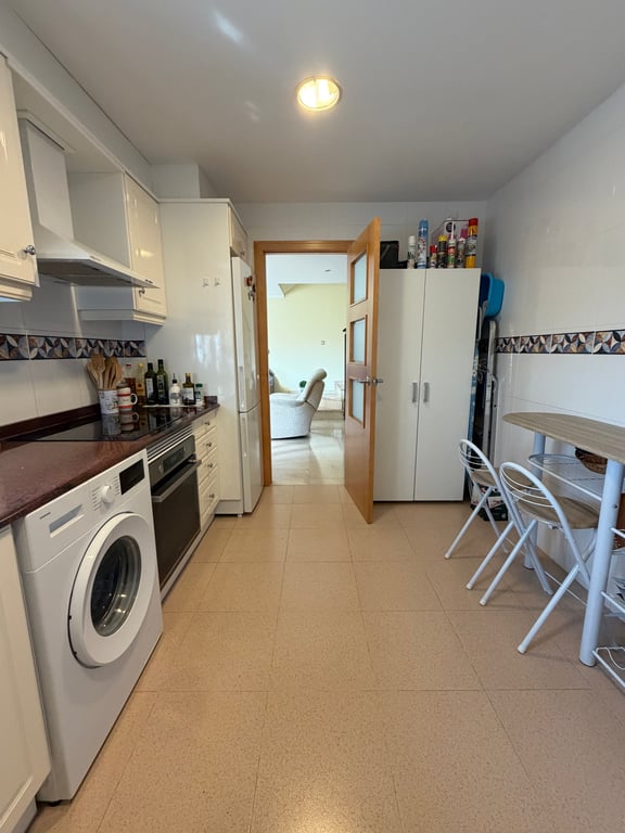 Cocina cerrada y equipada con armarios blancos brillantes, encimera oscura, lavadora y horno integrados, y una pequeña mesa de comedor. Una puerta de madera con cristal conecta con el salón. Cocina cerrada y equipada con armarios blancos brillantes, encimera oscura, lavadora y horno integrados, y una pequeña mesa de comedor. Una puerta de madera con cristal conecta con el salón.