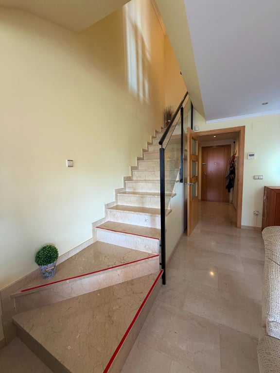 Escalera interior de mármol claro con barandilla moderna de metal negro y cristal, destacando las bandas antideslizantes rojas y el suelo a juego. La luz natural ilumina la pared de tono crema. Escalera interior de mármol claro con barandilla moderna de metal negro y cristal, destacando las bandas antideslizantes rojas y el suelo a juego. La luz natural ilumina la pared de tono crema.