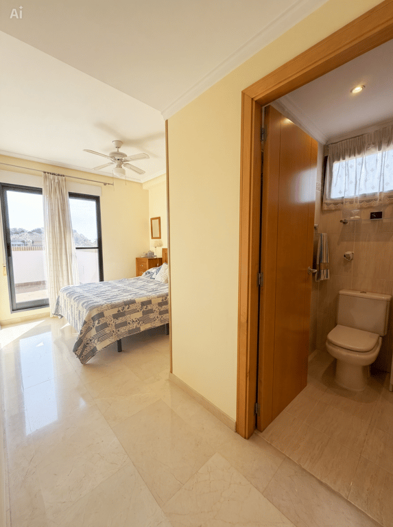 Luminoso dormitorio con suelos de mármol, cama doble y ventilador de techo, con acceso a terraza por puertas de cristal. Un baño alicatado con inodoro es visible parcialmente desde el pasillo. Luminoso dormitorio con suelos de mármol, cama doble y ventilador de techo, con acceso a terraza por puertas de cristal. Un baño alicatado con inodoro es visible parcialmente desde el pasillo.