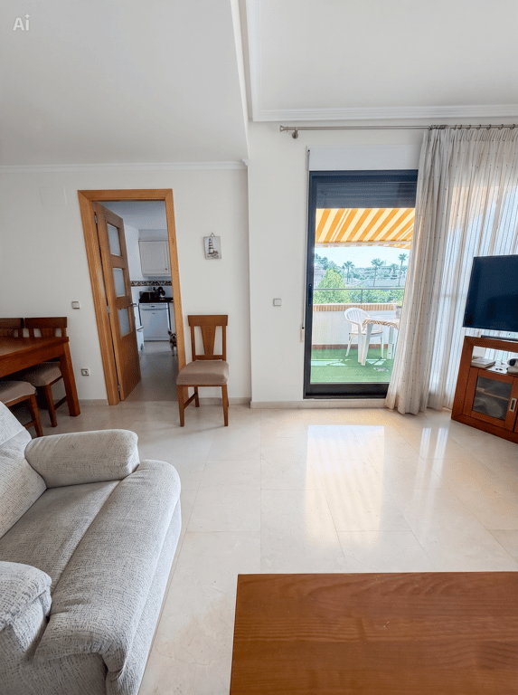Luminoso salón con suelo de baldosas claras, sofá y acceso a una terraza cubierta con césped artificial, mobiliario exterior y toldo rayado. Una puerta conduce a la cocina. Luminoso salón con suelo de baldosas claras, sofá y acceso a una terraza cubierta con césped artificial, mobiliario exterior y toldo rayado. Una puerta conduce a la cocina.