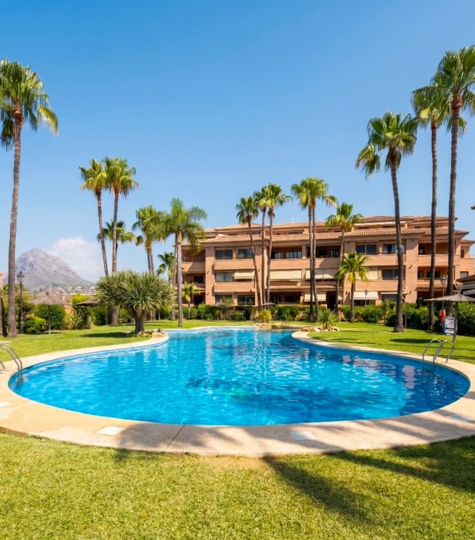 Vista de la piscina comunitaria de forma libre, rodeada de césped y palmeras, con el edificio residencial y el Montgó al fondo bajo un cielo despejado. Vista de la piscina comunitaria de forma libre, rodeada de césped y palmeras, con el edificio residencial y el Montgó al fondo bajo un cielo despejado.