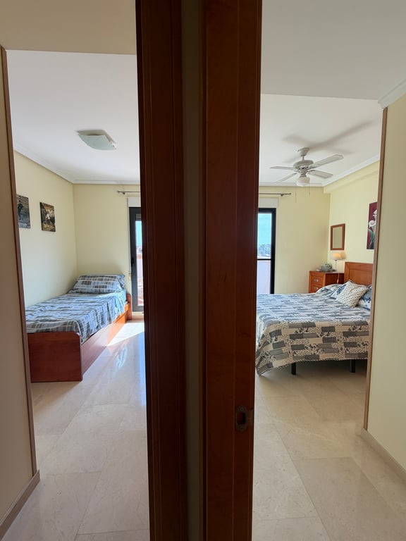 Vista desde el pasillo a dos dormitorios con suelos de mármol claro y paredes en tonos crema. El dormitorio principal incluye cama doble y ventilador de techo, mientras el segundo ofrece una cama individual. Vista desde el pasillo a dos dormitorios con suelos de mármol claro y paredes en tonos crema. El dormitorio principal incluye cama doble y ventilador de techo, mientras el segundo ofrece una cama individual.