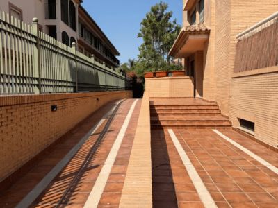 Acceso exterior con rampa de entrada al garaje subterráneo y escaleras a la entrada principal. Suelo de baldosas de terracota con franjas claras y muros de ladrillo.