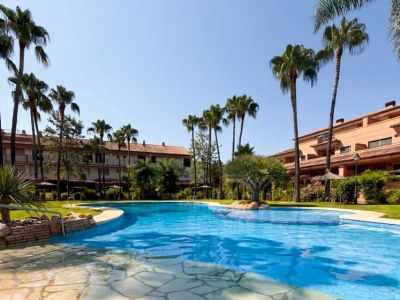 Amplia piscina de forma irregular con mosaicos azules, rodeada de pavimento de losas y césped verde. Palmeras altas y vegetación mediterránea enmarcan los edificios residenciales bajo un cielo azul claro.