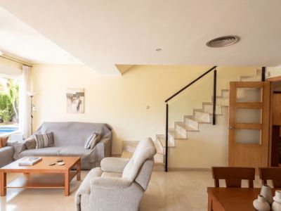 Amplia sala-comedor con suelo de baldosas claras y mesa de comedor de madera. Una escalera con barandilla negra conduce al piso superior, con una ventana que muestra la piscina exterior.