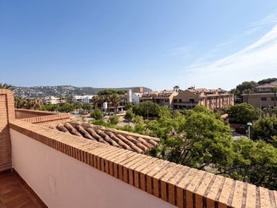 Amplia terraza con suelo de baldosas de terracota y barandilla de ladrillo. Ofrece vistas abiertas a la zona residencial, vegetación y colinas, con tejas antiguas visibles.