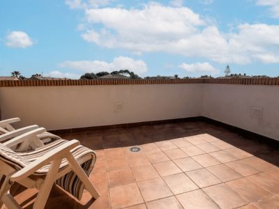 Amplia terraza exterior con suelo de baldosas de terracota, un murete blanco con remate de ladrillo y dos sillas plegables de madera, bajo un cielo azul con nubes.
