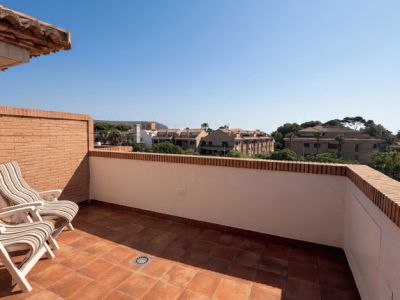 Amplia terraza exterior con suelo de terracota y paredes de ladrillo visto, equipada con dos sillas plegables. Ofrece vistas despejadas a la zona residencial y vegetación bajo un cielo azul.