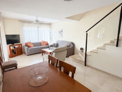 Amplio salón-comedor de planta abierta con mesa de madera, cómodos sofás y escalera moderna con barandilla de cristal. Aire acondicionado por conductos y suelos de baldosas.