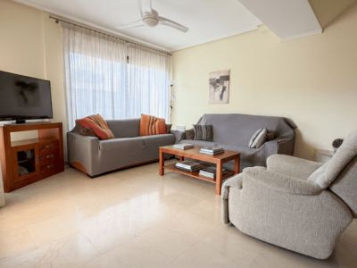 Amplio salón con suelo de baldosas claras efecto mármol, dos sofás confortables, un sillón reclinable y mesa de centro de madera. Luz natural a través del gran ventanal con cortinas translúcidas.