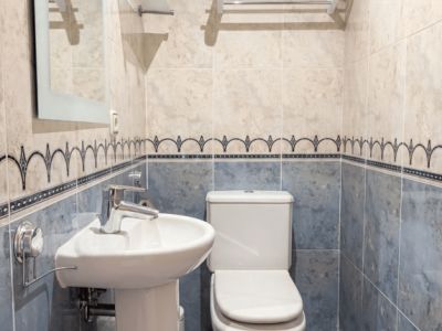 Aseo de cortesía compacto con lavabo de pedestal, inodoro y paredes alicatadas en dos tonos de azul y beige con cenefa decorativa. Incluye espejo y toalleros.