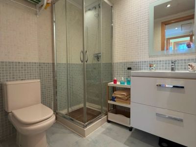 Baño funcional con ducha de cristal, inodoro en tono beige y un mueble de lavabo blanco con espejo iluminado. Las paredes combinan mosaicos claros y azules, complementados por el suelo de baldosas a juego.