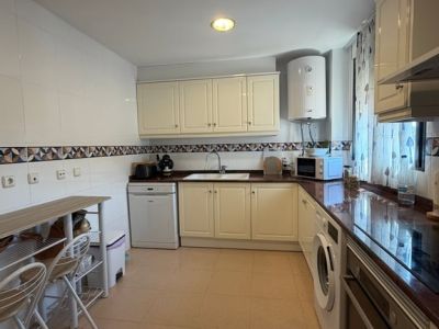 Cocina cerrada con armarios color crema, encimeras oscuras y electrodomésticos integrados. Incluye una barra de desayuno de madera y suelo de baldosas claras.
