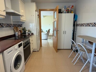 Cocina cerrada y equipada con armarios blancos brillantes, encimera oscura, lavadora y horno integrados, y una pequeña mesa de comedor. Una puerta de madera con cristal conecta con el salón.
