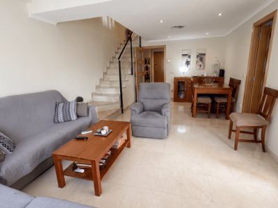 El salón-comedor de planta abierta presenta suelos de baldosas claras, un cómodo sofá gris, una mesa de centro de madera y un conjunto de comedor. Una escalera de mármol con barandilla negra conduce al nivel superior.