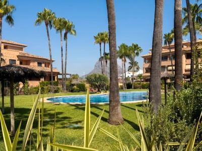 La piscina comunitaria está rodeada por césped verde y altas palmeras, creando un oasis tropical con sombrillas y edificios residenciales al fondo bajo un cielo azul claro.