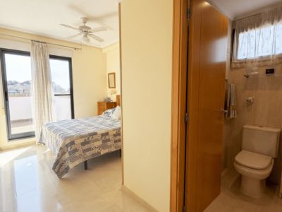 Luminoso dormitorio con suelos de mármol, cama doble y ventilador de techo, con acceso a terraza por puertas de cristal. Un baño alicatado con inodoro es visible parcialmente desde el pasillo.