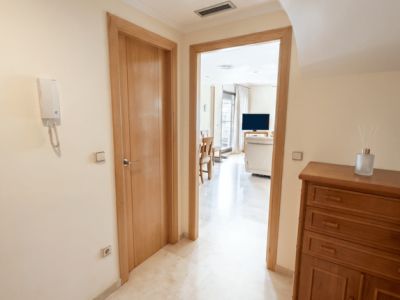 Luminoso recibidor con suelo de mármol pulido y puertas de madera clara. Un mueble auxiliar de madera se sitúa a la derecha, con vistas al salón-comedor.