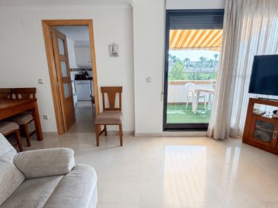 Luminoso salón con suelo de baldosas claras, sofá y acceso a una terraza cubierta con césped artificial, mobiliario exterior y toldo rayado. Una puerta conduce a la cocina.