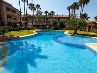 Piscina comunitaria de agua salada con mosaicos azules, rodeada de césped, palmeras y edificios residenciales bajo un cielo despejado.