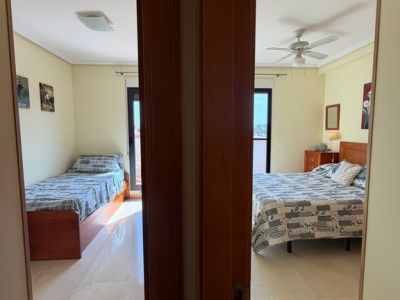 Vista desde el pasillo a dos dormitorios con suelos de mármol claro y paredes en tonos crema. El dormitorio principal incluye cama doble y ventilador de techo, mientras el segundo ofrece una cama individual.