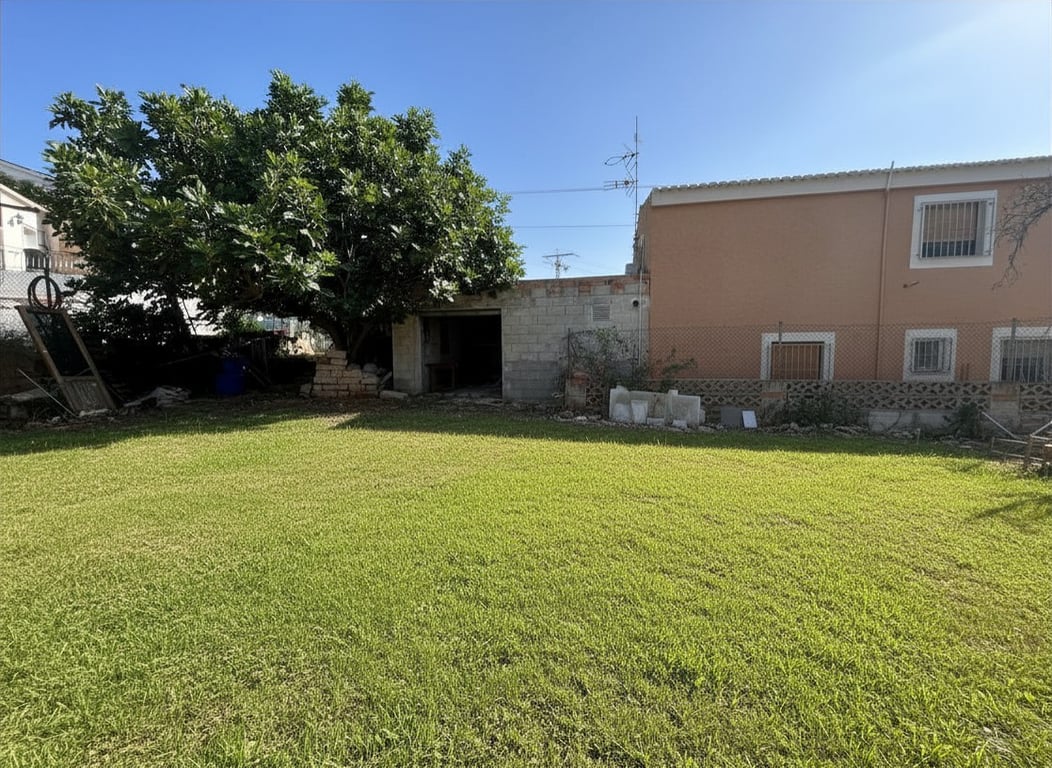 Amplio jardín privado con césped verde, un gran árbol frondoso y una edificación auxiliar de bloques junto a la fachada principal color melocotón. Amplio jardín privado con césped verde, un gran árbol frondoso y una edificación auxiliar de bloques junto a la fachada principal color melocotón.