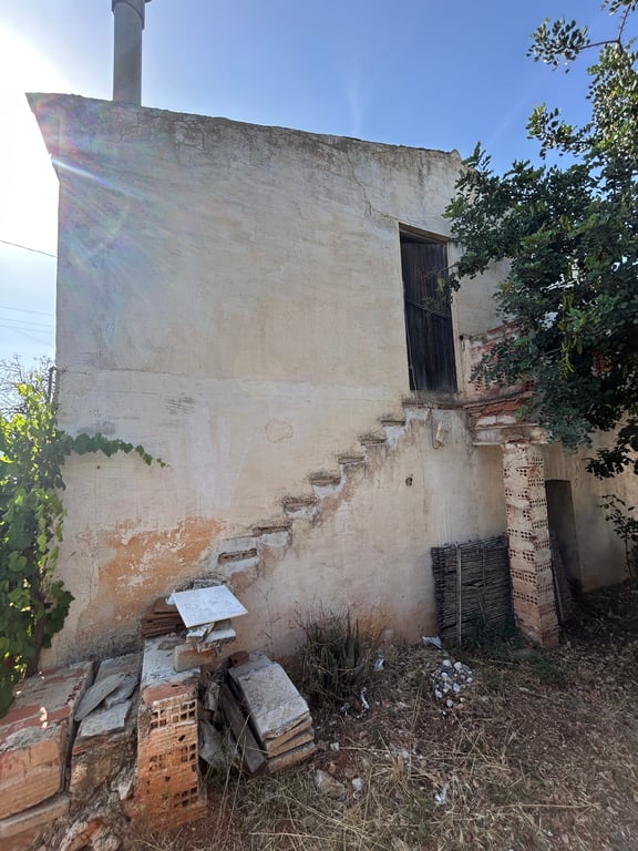 Exterior de la propiedad con fachada enlucida y agrietada, escalera exterior incompleta y puerta de madera en el nivel superior, rodeado de escombros y vegetación. Exterior de la propiedad con fachada enlucida y agrietada, escalera exterior incompleta y puerta de madera en el nivel superior, rodeado de escombros y vegetación.