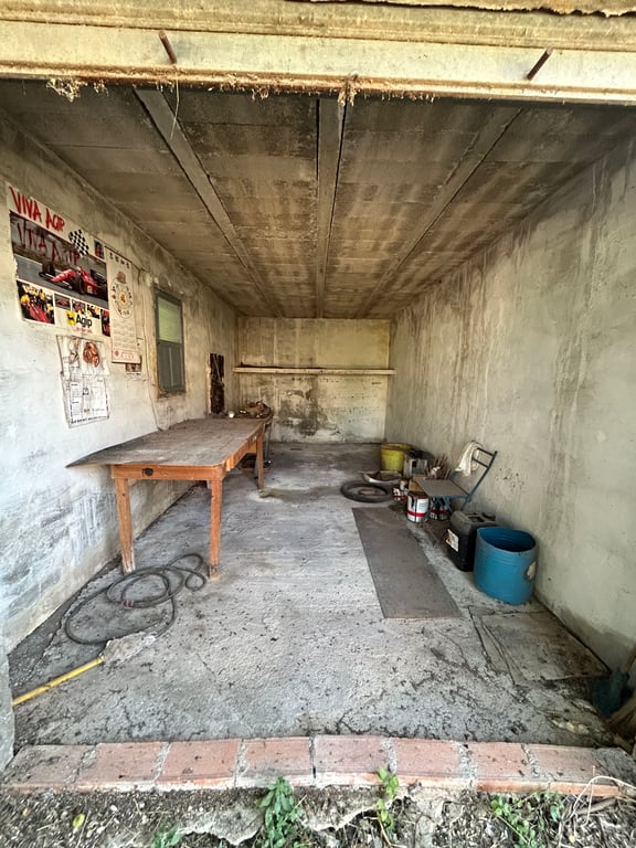Interior de un garaje o almacén con paredes y techo de hormigón visto. Incluye una mesa de madera, una ventana y objetos variados, mostrando un estado que requiere reforma. Interior de un garaje o almacén con paredes y techo de hormigón visto. Incluye una mesa de madera, una ventana y objetos variados, mostrando un estado que requiere reforma.