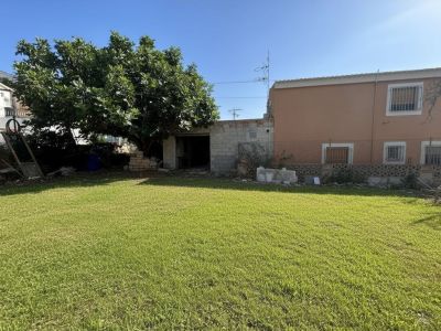 Amplio jardín privado con césped verde, un gran árbol frondoso y una edificación auxiliar de bloques junto a la fachada principal color melocotón.