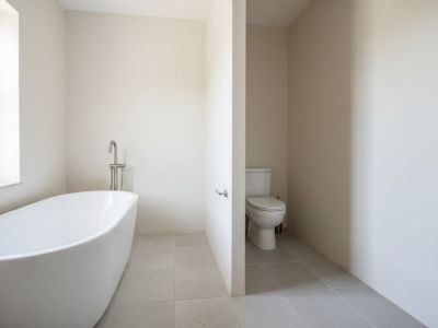 Baño con bañera exenta blanca y grifo de pie, suelo de baldosas grises y ventana. El inodoro se ubica en una alcoba separada por una pared parcial.