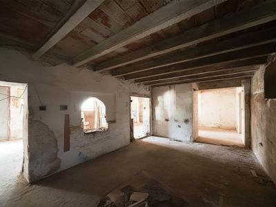 Estancia interior en estado de reforma, con vigas de madera expuestas en el techo, paredes blancas rugosas y múltiples accesos a otras habitaciones.
