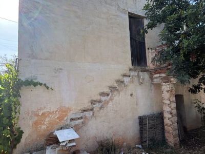 Exterior de la propiedad con fachada enlucida y agrietada, escalera exterior incompleta y puerta de madera en el nivel superior, rodeado de escombros y vegetación.