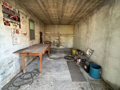 Interior de un garaje o almacén con paredes y techo de hormigón visto. Incluye una mesa de madera, una ventana y objetos variados, mostrando un estado que requiere reforma.