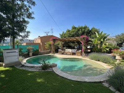 Piscina de forma irregular con borde de piedra junto a una pérgola con buganvillas rosas, creando una zona de estar exterior sombreada en un jardín mediterráneo.